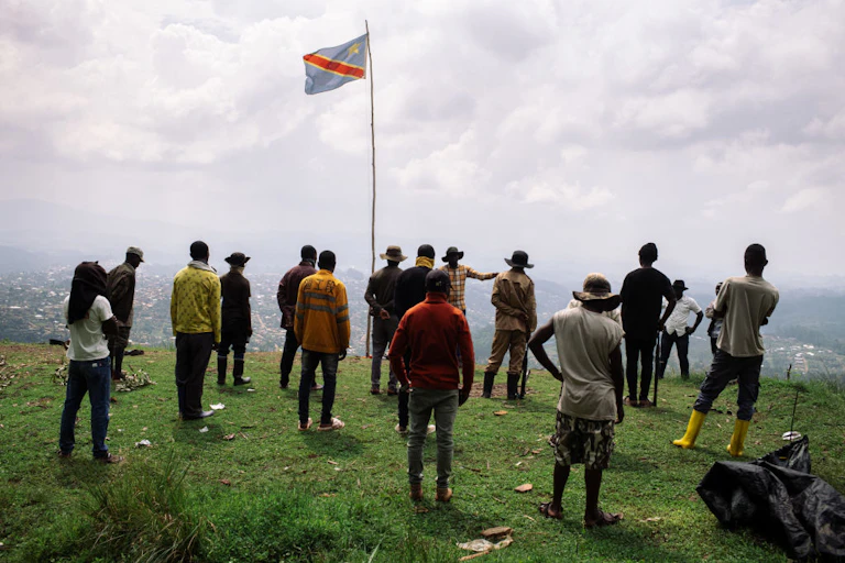 A group of standing men face a speaker standing on a hill overlooking a town below with the Congolese flag fluttering above them