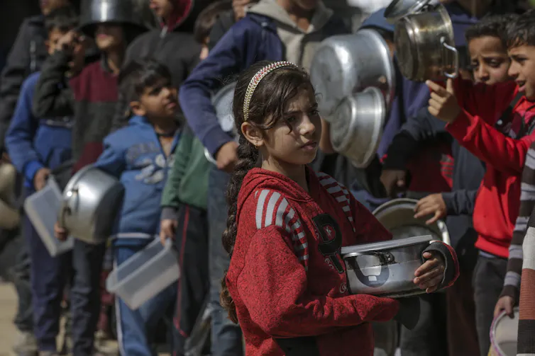 A little girl stands in a queue in GAza holding an empty bowl.