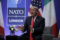 A man in a suit with blond-grey hair gestures as he speaks with a NATO sign behind him.