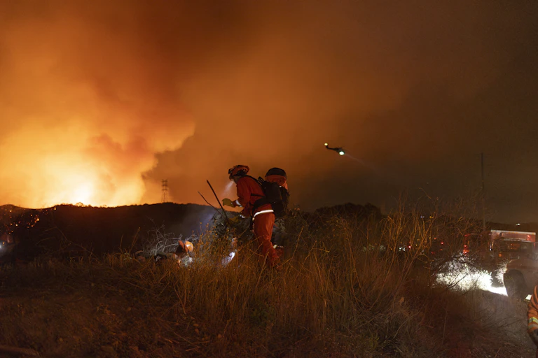 Four firefighters in a brushy area with flames rising into an orange sky
