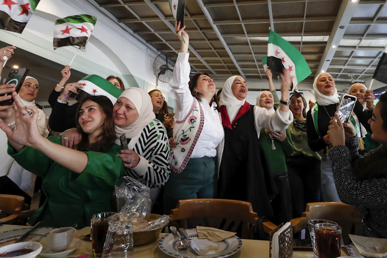 A group of women smile and celebrate, waving Syrian flags.