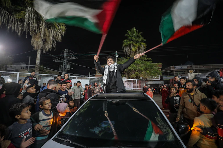 A young manstanding in a car waves Palestinian flags as people around him celebrate.