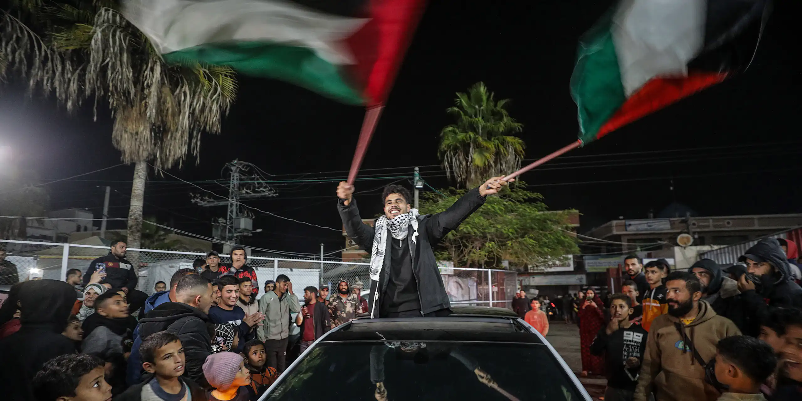 A young manstanding in a car waves Palestinian flags as people around him celebrate.