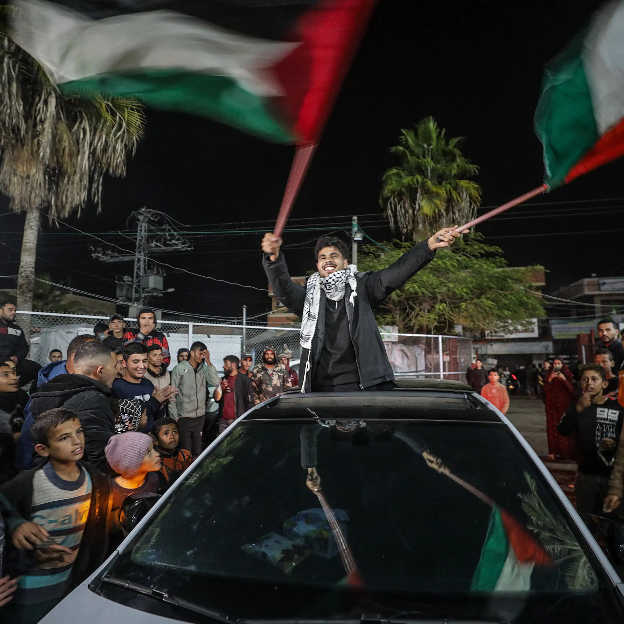 A young manstanding in a car waves Palestinian flags as people around him celebrate.
