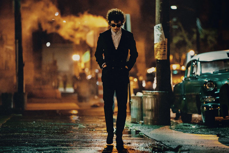 A lone male figure in sunglass and skinny suit walking down a 1960s NYC street at night
