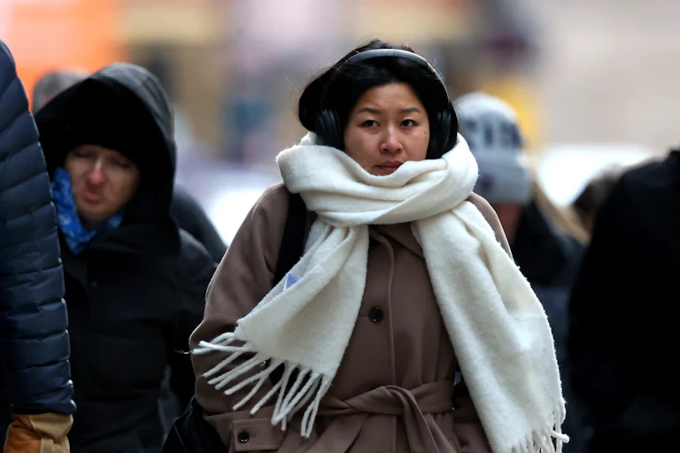 A woman wrapped in a thick scarf and ear muffs walks with people with thick gloves and hoods on a clearly chilly day.