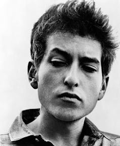 A portrait of a young Bob Dylan with sticky up hair and a pensive look in black and white.