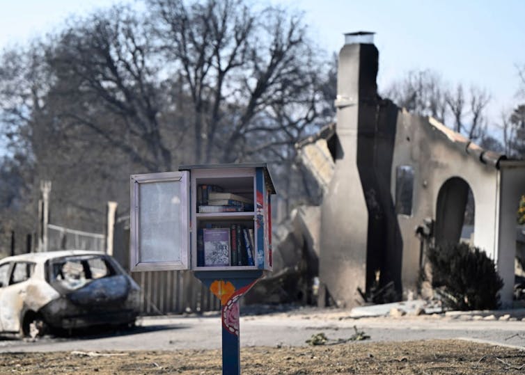 A container for books with its door open sits on a post in front of a scorched car and the rubble of a home.