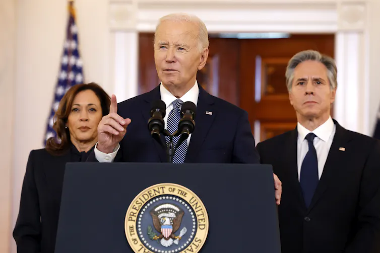 A white man with a dark suit stands at a podium and raises a finger. Two people, one man and a woman, stand behind him, alongside an American flag.