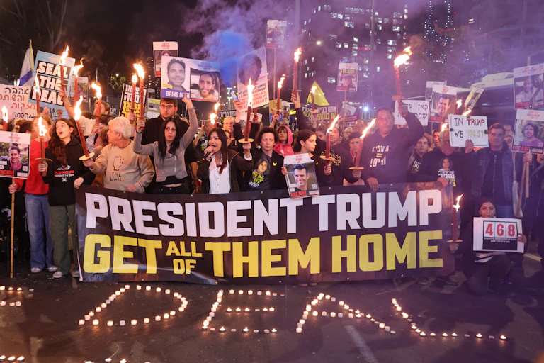 A crowd of people hold torches and signs and stand in front of a banner that says 'President Trump get all of them home.'