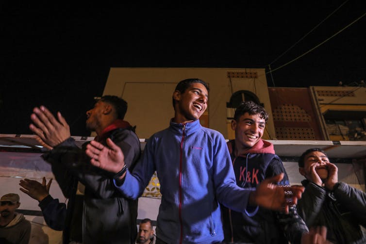 Three young men are seen waving their hands and clapping and smiling at nighttime.