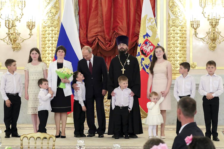 Nine children of various ages stand on an ornate platform with a woman and two men, with the Russian flag in the background.