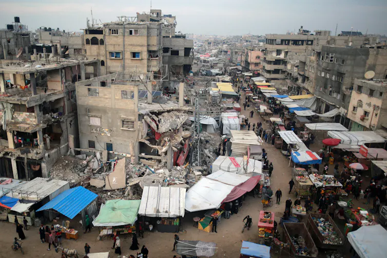 Many destroyed buildings are seen on a gray day. People walk past outside stalls on a street.