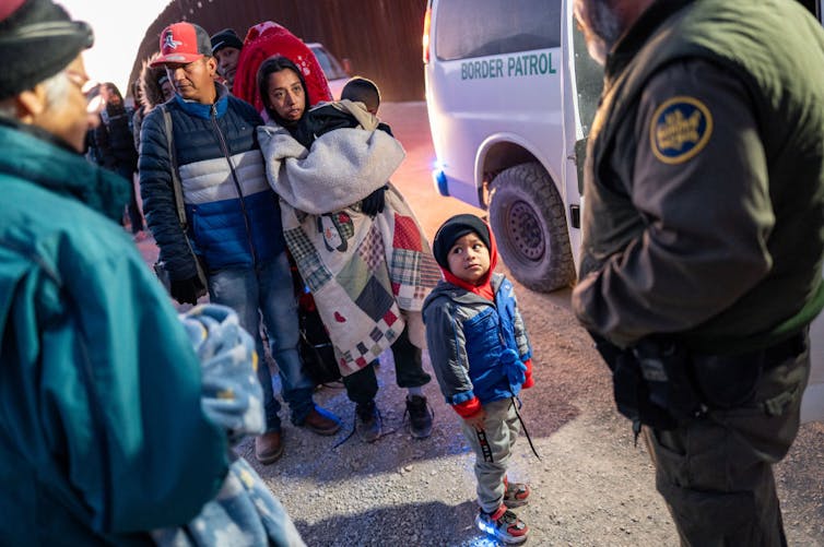 A small boy wearing a jacket stands in front of a woman covered in a blanket and holding a baby. Other people, including a man with a green jacket, stand nearby and next to a white van.