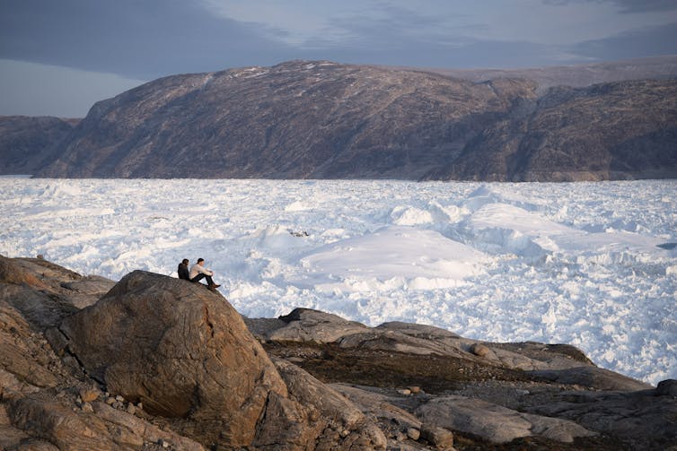 Two people sit on a ridge overlooking a large glacier.