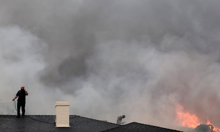 Two people stand on rooftops with garden hoses as a home burns nearby.