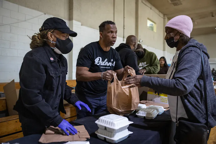 A man standing behind a table with boxes of food hands a bag to a woman in line.