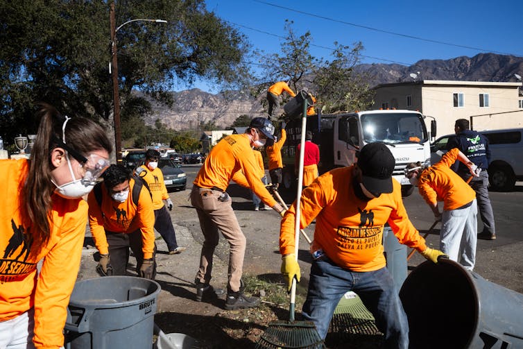 Several people wearing bright orange shirts rake debris into garbage cans on a residential street.