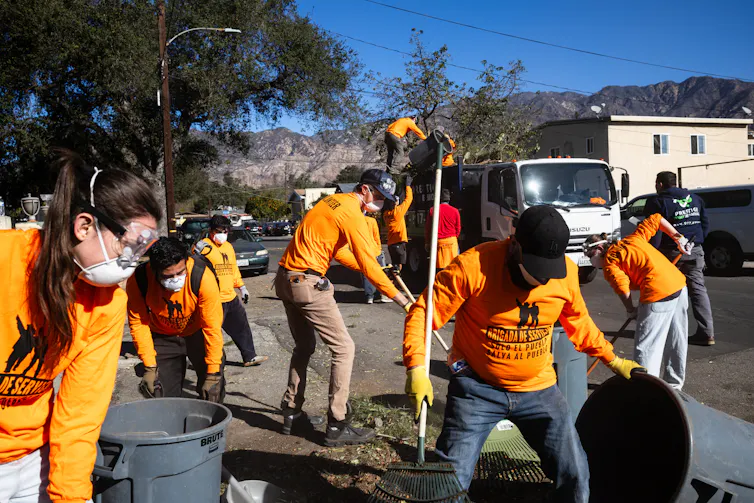 Several people wearing bright orange shirts rake debris into garbage cans on a residential street.