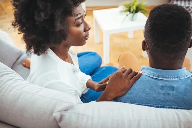 A serious-looking woman touches a man's shoulder as they sit on a couch.