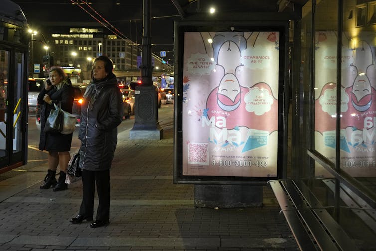 Two women stand near an outdoor bus stop on a street with a broad sidewalk at night.