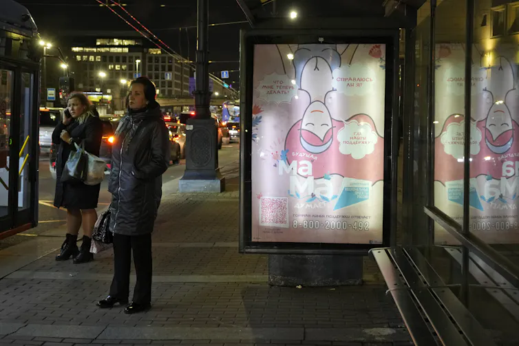 Two women stand near an outdoor bus stop on a street with a broad sidewalk at night.