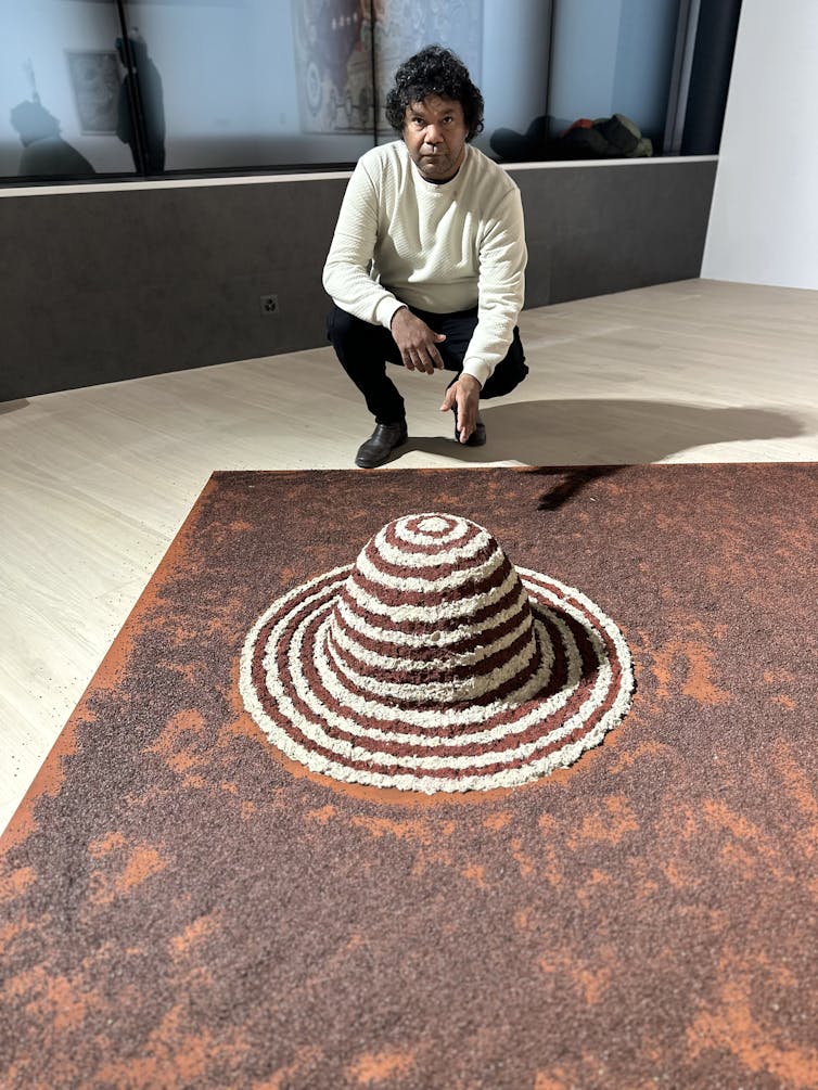A man kneels over his artwork: a small red mound with white concentric circles.