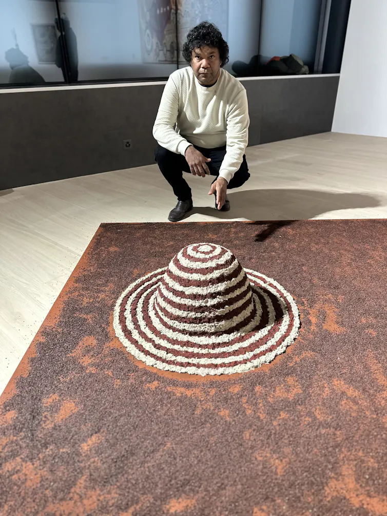 A man kneels over his artwork: a small red mound with white concentric circles.