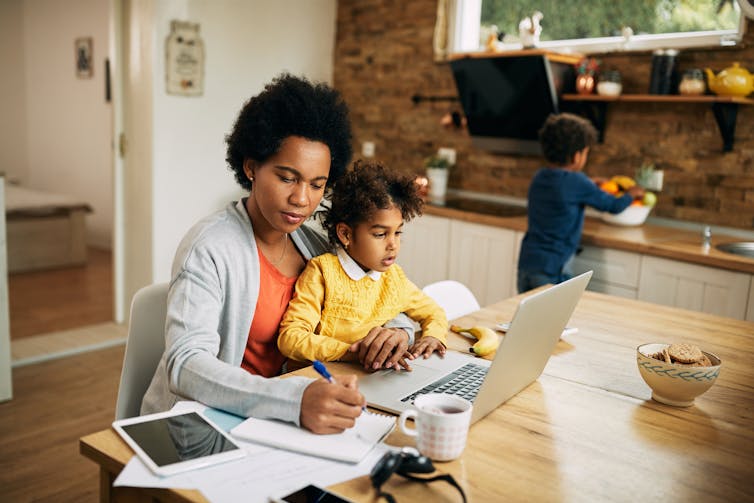 Working mother taking notes while daughter is sitting on her lap and using laptop
