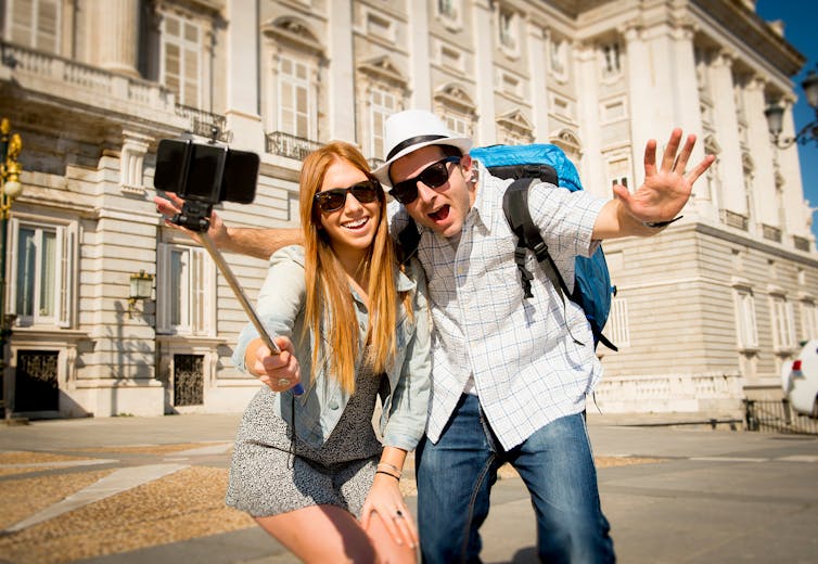 A young couple in sunglasses and backpacks poses with a selfie stick in front of a grand building