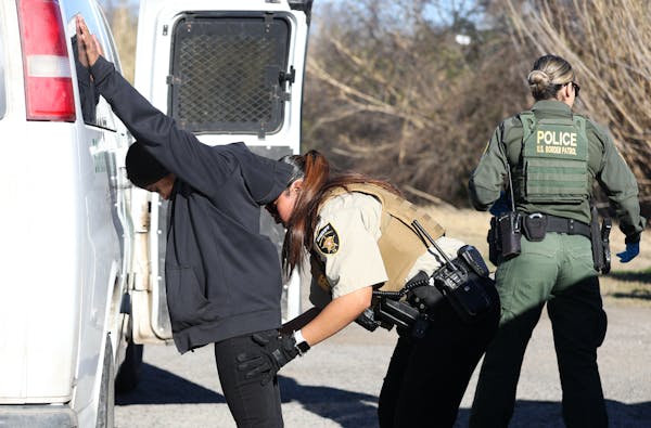 A woman in a uniform searches a man dressed in black who is leaning against a van outside.