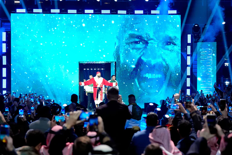Tyson Fury in red gown in front of screen bearing his face.