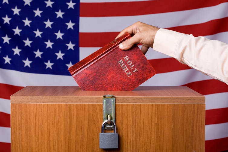 A human hand inserting the Bible into a locked ballot box, placed in front of the American flag.