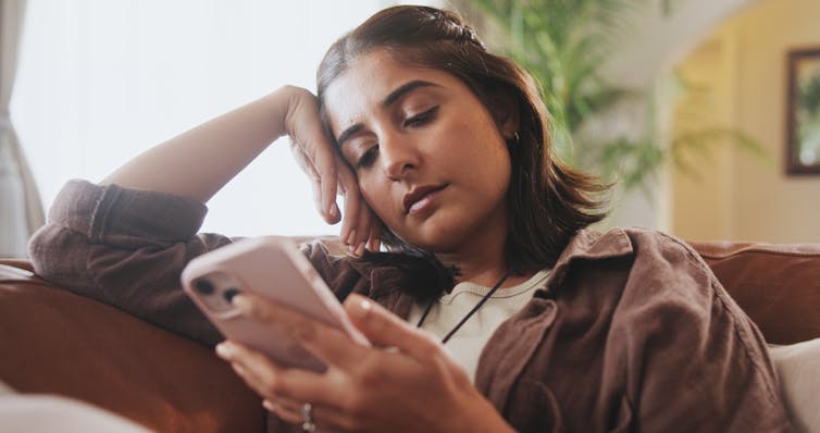 A young woman relaxes on a sofa, scrolling listlessly on her mobile phone
