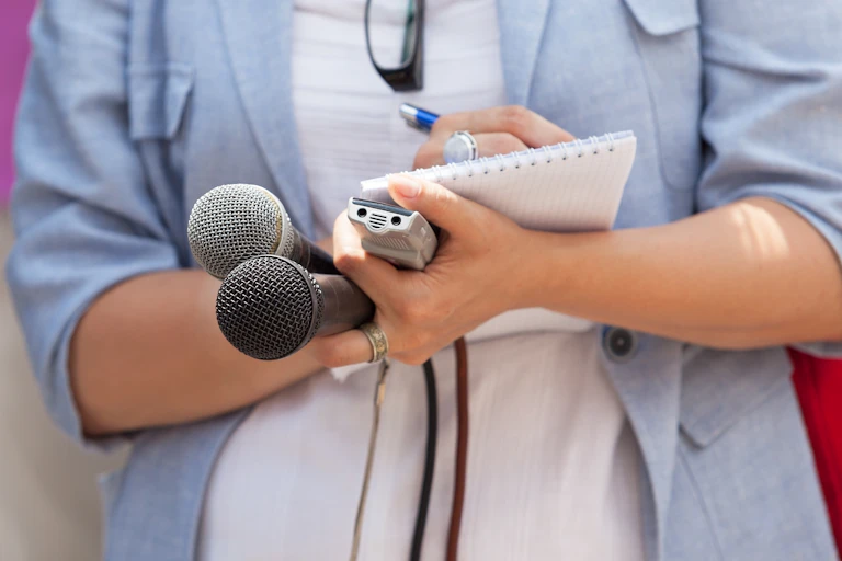 A journalist's hands full with two microphones and an audio recorder while she writes with a pen on a paper notepad