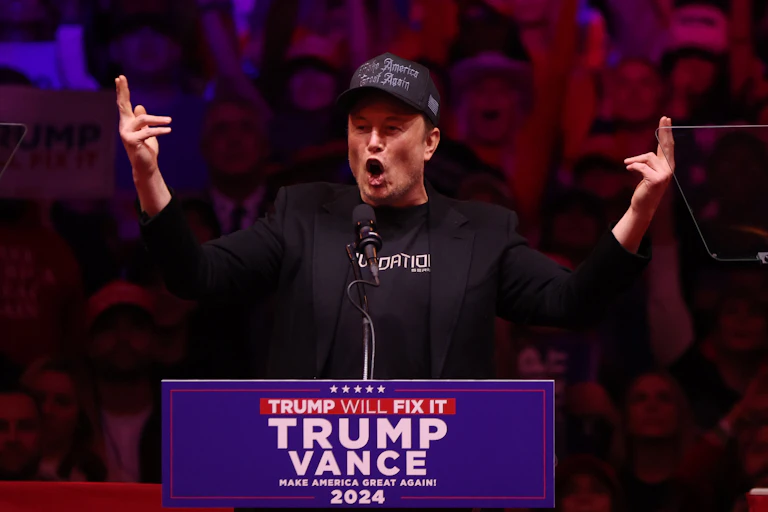 Elon Musk speaks to a crowd wearing a Make America Great Again cap