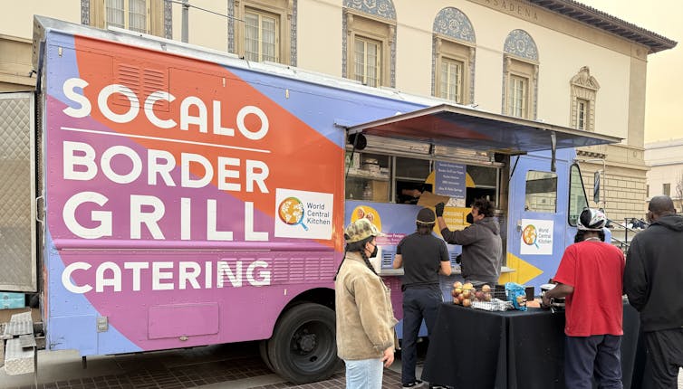 People line up outside of a food truck that reads "Socalo Border Grill Catering".
