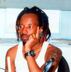 A young African man with dreadlocks sits in a white vest, his face resting on his hand.
