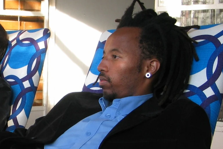 A young African man with dreadlocks reclines in a chair.
