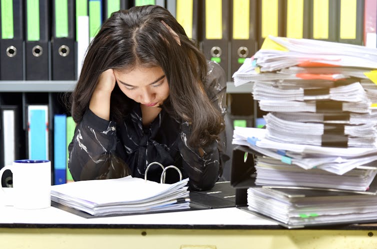 Seated woman holding her head with large pile of files beside her