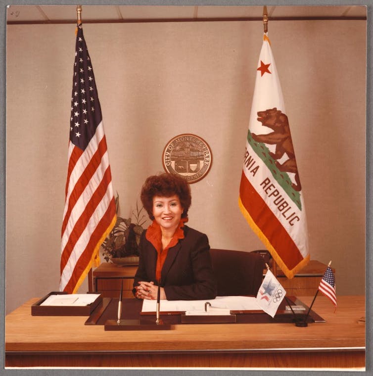 A photograph from the 1980s of a professionally dressed woman sitting behind a desk flanked by the American flag and the flag of the state of California.