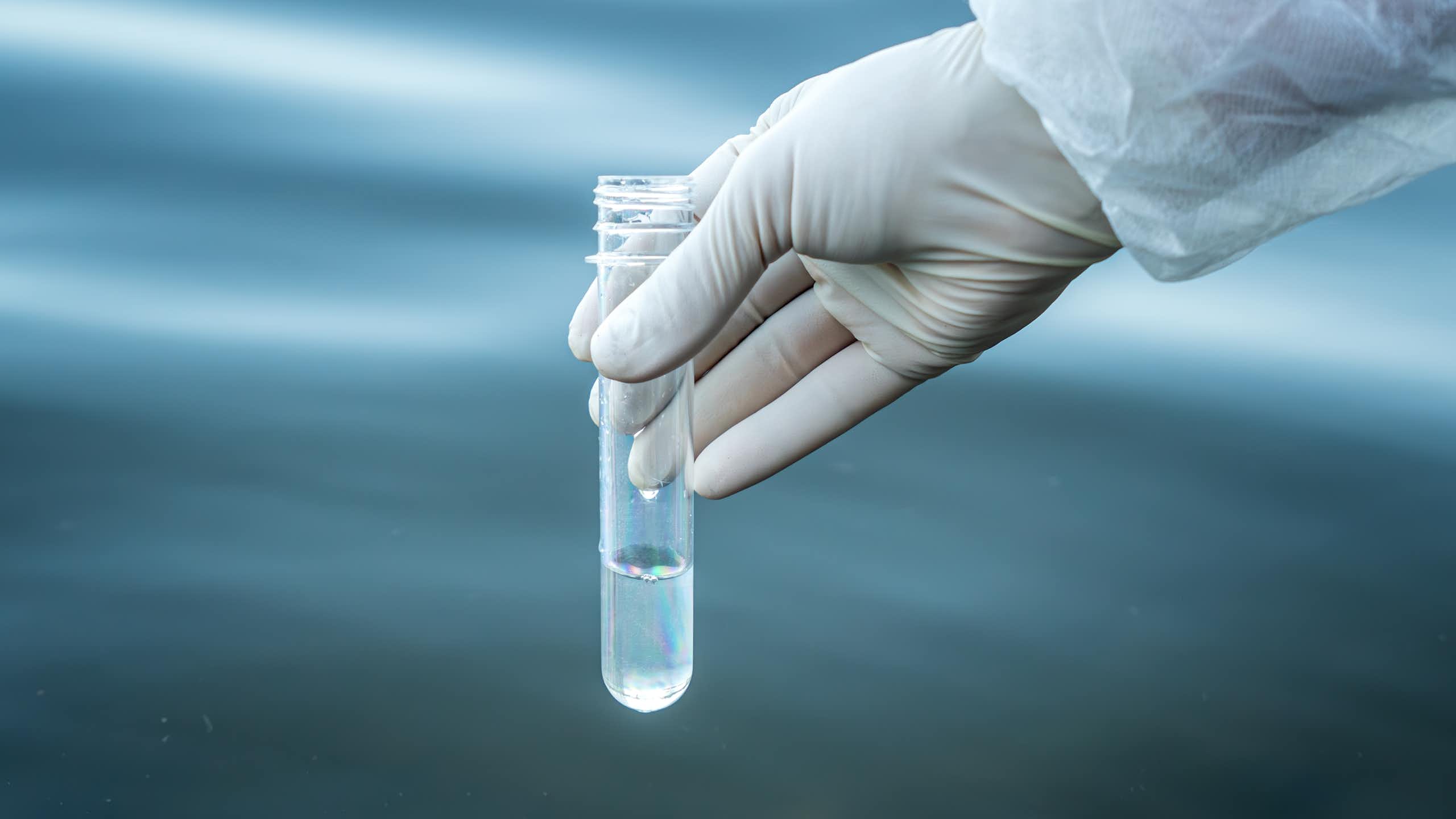 scientist hand in white latex glove and labcoat holds testtube of water, water in background