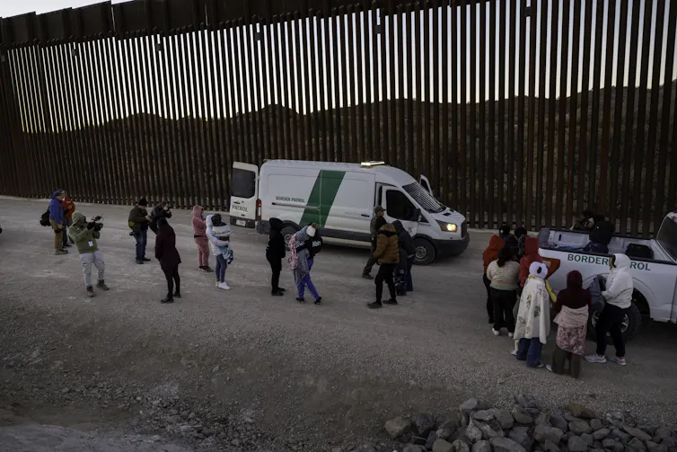 A small group of people are seen in front of a large dark wall with slits in it. Two white-and-green vans are parked next to the wall.