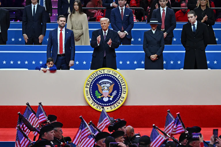 A man wearing a jacket stands and slaps and is surrounded by other people who are dressed formally and also standing. People in a band hold US flags below them.