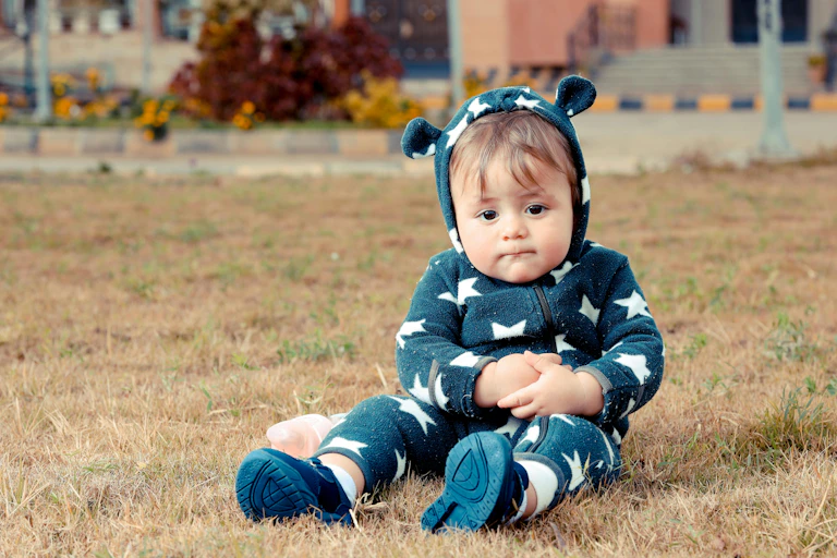 A cute child in a fuzzy onesie sits on grass outdoors.