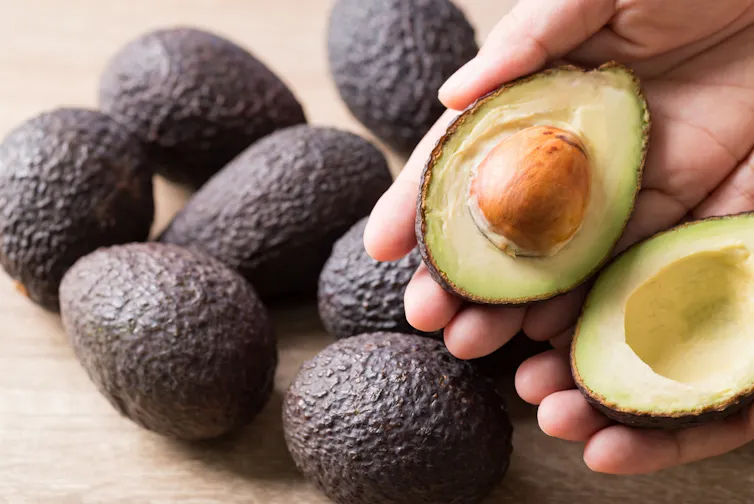 A pile of dark avocados on a cutting board with one cut open in the foreground.