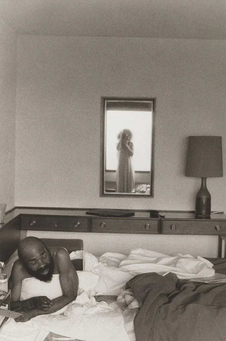 A man in a bed, the photographer in a mirror.