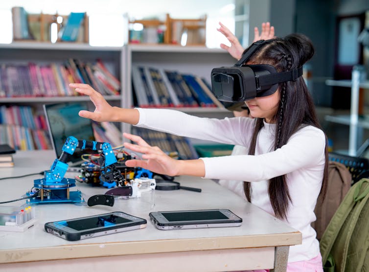 Two young students with VR glasses in a room full of books, various types of tools and robotic arms.