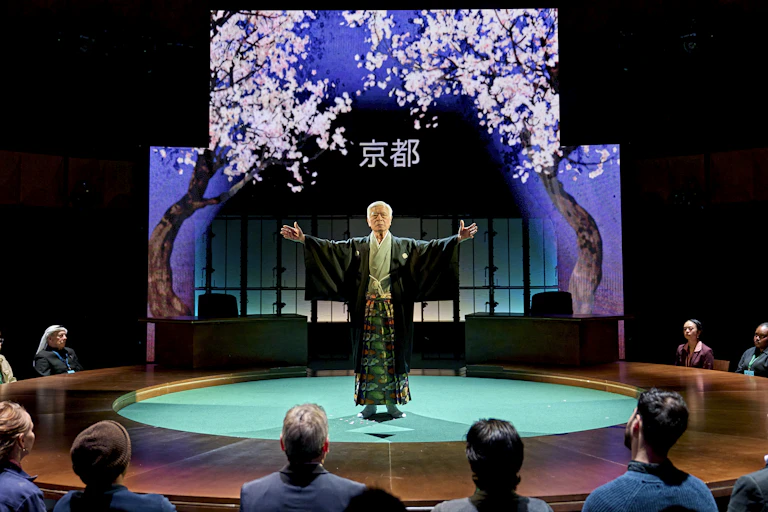 A Japanese man in traditional kimono dress standing on a stage with cherry blossom framing his figure.