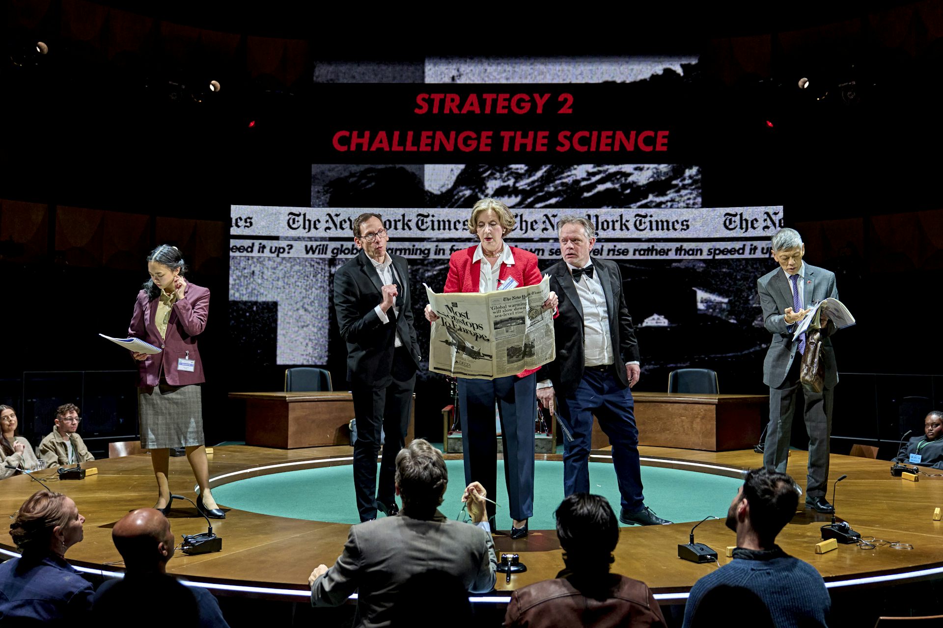 A group of actors on a stage with a supertitle above them saying: Challenge the science.